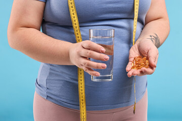 Plus size woman holding pile of weight loss supplements and glass with water on light blue background, closeup