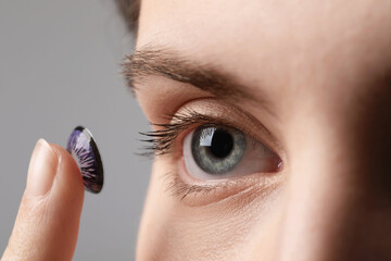 Woman putting in color contact lens on grey background, closeup