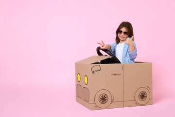 Little girl showing thumbs up while driving car made with cardboard on light pink background. Space...