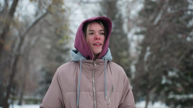 Lady jogging in cold winter park with thoughtful facial expression, dressed in warm hoodie and coat, snow-covered trees in background
