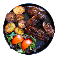 Black ceramic plate contains furnace baked pork ribs, fried potatoes and pieces of fresh vegetables. Meat is complemented with tomato slices and arugula leaves. Isolated over white background