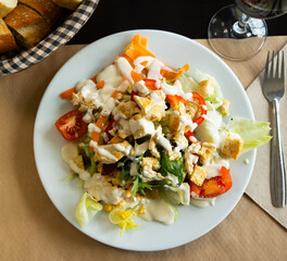 Caesar salad with slices of fried chicken, fresh tomatoes, corn and olives on a white plate. Delicious serving of meal with vegetables and meat. Lunch option in a café
