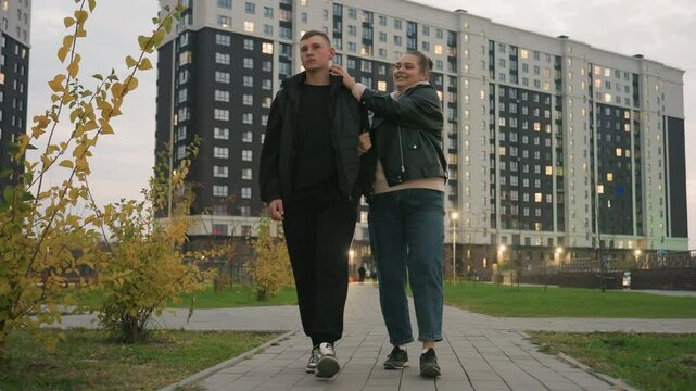 Romantic moment as couple walk along paved path in urban park while smiling lady playfully tickles her partner who tries to tickle her back but she gently holds his hand teasingly with affection