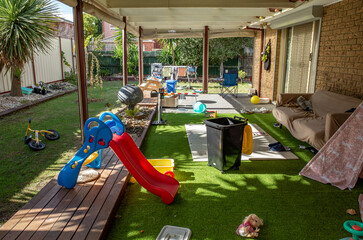 A cluttered and messy backyard play area featuring artificial green grass, a toddler's slide, and...