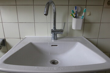 Close-up of a bathroom sink with a chrome faucet and water running slowly. Toothbrushes in a wall-mounted holder. Daily hygiene and water use in a simple home bathroom setting