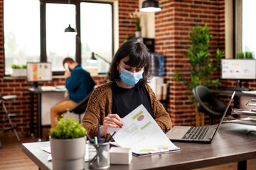 Businesswoman seated at tidy desk with laptop, examining detailed financial documents in brick wall...