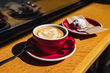 Red coffee cup with creamy latte art sits in golden sunlight beside cookie on napkin. Coffee moment feels warm and inviting with cozy atmosphere and perfect coffee pairing