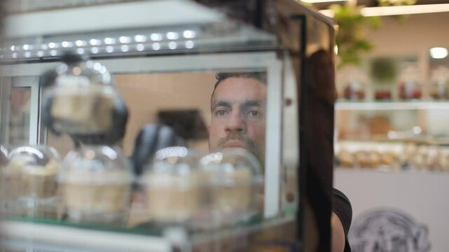 Focused hispanic owner of a small takeaway shop organizing cheesecake cups in a display fridge. Reflected through glass, showing the calm rhythm of a local food business.