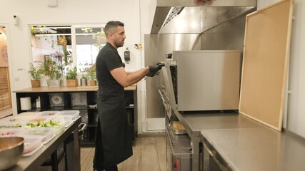 Owner or chef in a takeaway shop puts a tray of food into the oven. A moment of everyday prep inside a cozy, professional kitchen filled with fresh ingredients and plants.
