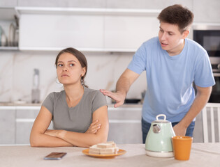 Annoyed young girl sitting sadly while guy is brawling with her in the kitchen