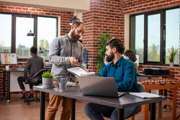 Caucasian man sits with laptop as male coworker checks clipboard and explains updates on market analysis. Bearded businessmen are actively collaborating at office desk, planning project presentation.