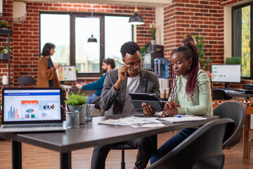 Colleagues share marketing concepts, collaborating with a tablet in modern workspace. African American researchers engage in intense dialogue, analyzing strategies and reviewing plans on device.