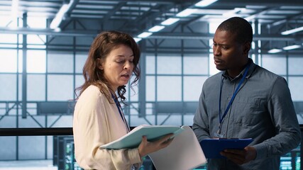 Fototapeta premium Cheerful admins meeting in data center to review reports and files, doing handshake. Server room employee saluting colleague, doing troubleshooting to prevent downtime, camera A