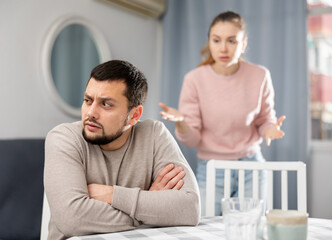 Offended young man sitting at table at home with disgruntled woman behind his back. Couple relationship problems concept..