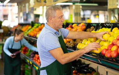 Focused elderly male supermarket worker in apron arranging Golden Delicious apples in fruit section...