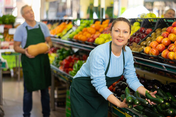 Positive middle-aged woman, skilled market assistant arranging organic cucumber on shelves in hypermarket
