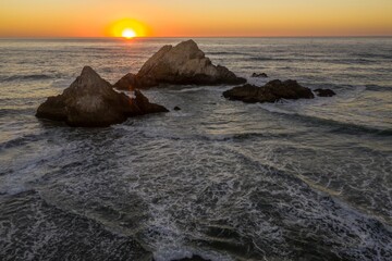 Sunset over the Pacific Ocean inOcean Beach, San Francisco, California, USA. The sun is setting behind large rocks in the ocean, creating a beautiful and serene scene.