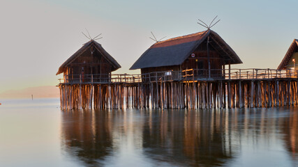 Obraz premium Wooden stilt houses stand over calm water at sunrise in a serene lakeside location with reflections and gentle light