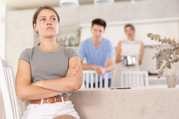 Obraz premium Sad offended young woman sitting at kitchen table during argument with dissatisfied husband and mother-in-law standing behind on blurred background. Concept of family fights