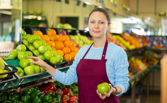 Skillful female merchandiser in uniform checking green apples before selling in grocery supermarket