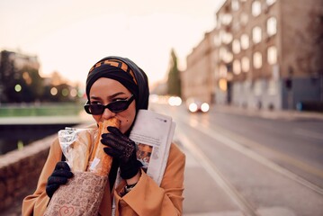 Obraz premium A beautiful Arab woman, elegantly dressed in a modern hijab, confidently walks through the city streets, carrying a handbag and newspaper.