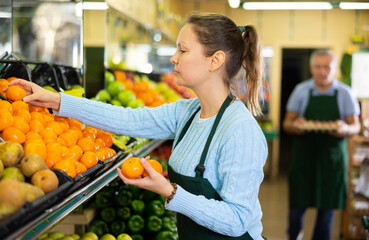 Positive middle-aged saleswoman setting out tangerines on food stall in grocery store