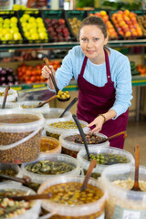 Cheerful middle-aged woman pleasantly offering olives in scoop in large vegetable shop