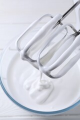 Mixing whipped cream with hand mixer in bowl on white wooden table, top view