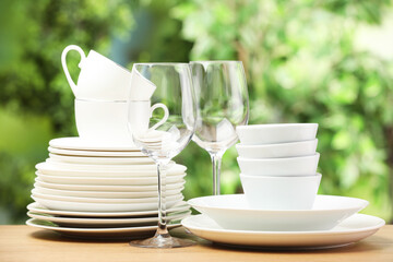 Different clean dishware, cups and glasses on wooden table against blurred green background