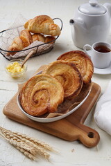 A delicious breakfast spread featuring swirl pastries and croissants, served with a cup of tea, butter, and a white teapot on a rustic wooden board, creating a warm and cozy morning vibe.