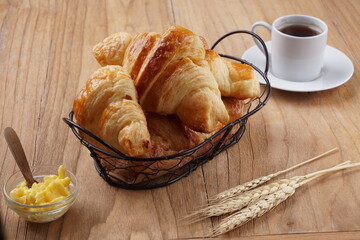 A basket of freshly baked croissants placed on a wooden table, accompanied by a glass bowl of butter, a white cup of black coffee, and wheat stalks, creating a warm and inviting breakfast scene.