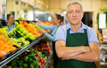 Positive aged salesman standing in front of food stall in grocery store with large assortment