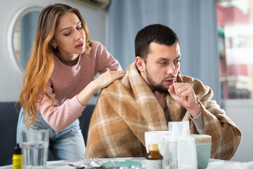 Woman comforting her ill husband who sitting at table with medicines at home.