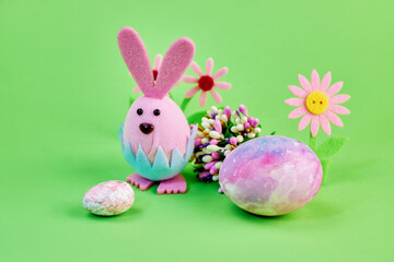 cute toy pink bunny celebrates Easter with big and small colorful eggs and flowers on a green background