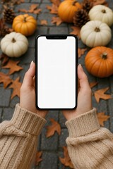 Mockup of a smartphone with blank white screen held by a woman wearing a beige knitted sweater. The autumn-themed background features pumpkins, pinecones, and fallen leaves on a stone path.