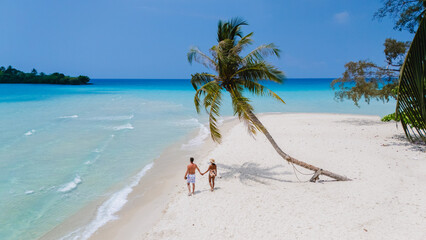 A couple strolls hand in hand across the soft, white sands of Koh Kood Island Thailand, perfectly nestled among stunning turquoise waters and lush palm trees, crafting a breathtaking paradise scene