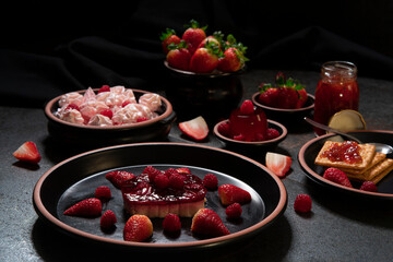 Strawberry dessert with raspberries on a black ceramic plate, soda crackers with strawberry jam and strawberry jelly, strawberry candies and a pot of strawberries on a black table 