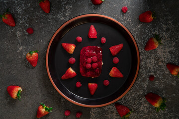 Top view of strawberry and raspberry dessert on a round black ceramic plate surrounded by strawberries