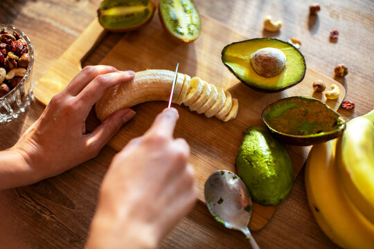 Woman slicing banana for smoothie with fresh avocado and nuts on kitchen table