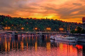 Fototapeta premium The elegant Svatopluk Čech Bridge gracefully spans the Vltava River in Prague, Czech Republic, at twilight, with its lights mirroring on the water alongside docked tourist boats.