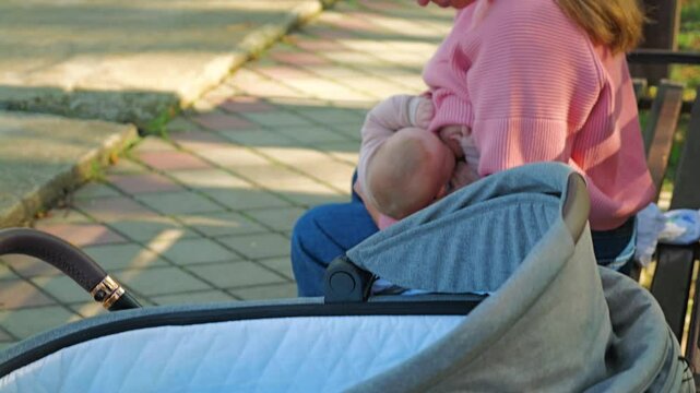 A woman wearing glasses and a pink jumper breastfeeding a newborn baby sitting in an outdoor park. In the foreground is a baby pram with a carrycot. Serene nature of autumn park with bright foliage