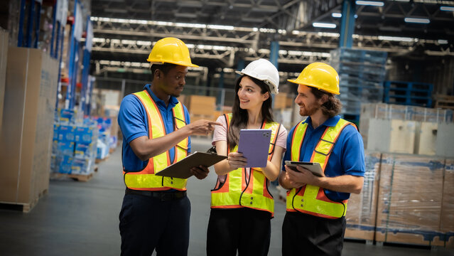 Three workers in safety vests are standing in a warehouse, looking at a tablet. They seem to be discussing something important