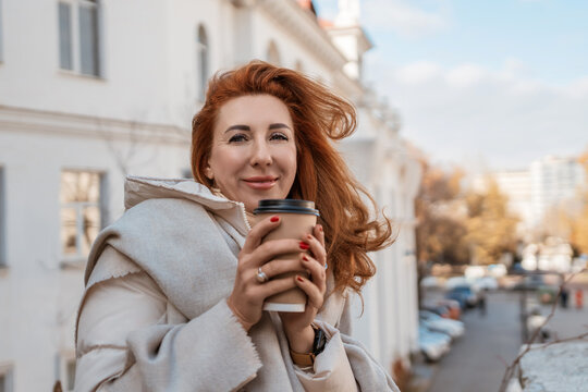Woman Coffee CityStreet Autumn: Redhead enjoys coffee outdoors on a windy autumn day near European-style buildings.