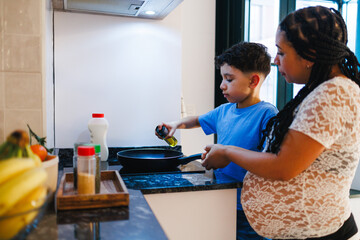 Latin pregnant mom helping her son pouring olive oil into frying pan in modern kitchen