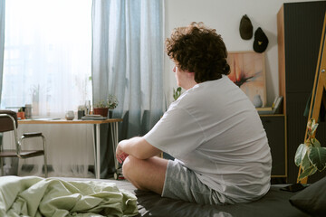 Portrait of a man sitting on bed in sunlit room, looking out window, surrounded by indoor plants...