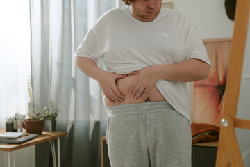 Obraz premium Man examining his stomach in casual home setting with natural light filtering through the curtains. Room features cozy decor with a small table and indoor plants