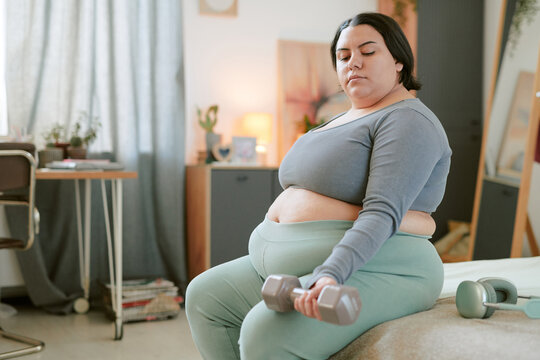 Person sitting on edge of bed holding dumbbell and exercising in bedroom setting with furniture in background