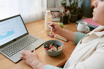Person engaged in multitasking by eating breakfast while working on laptop at home. Various personal items and healthy food options are visible on desk