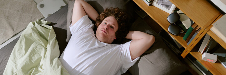 Portrait of person lying on bed with arms behind head, surrounded by books and electronics,...
