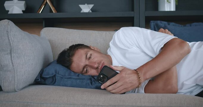 Sad man lies on the bed and looks at his smartphone. Depressed man, suffering from a breakup with his girlfriend, sad male lies on couch and waits for a message from his girlfriend. Breakup concept